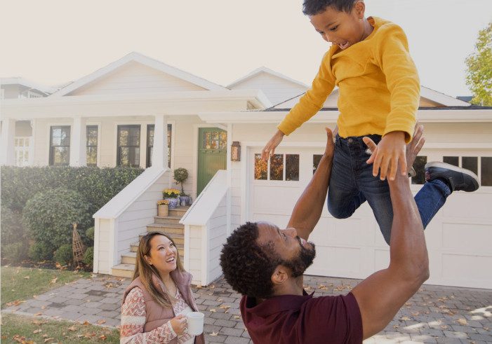 Man lifts child with woman, house in background.