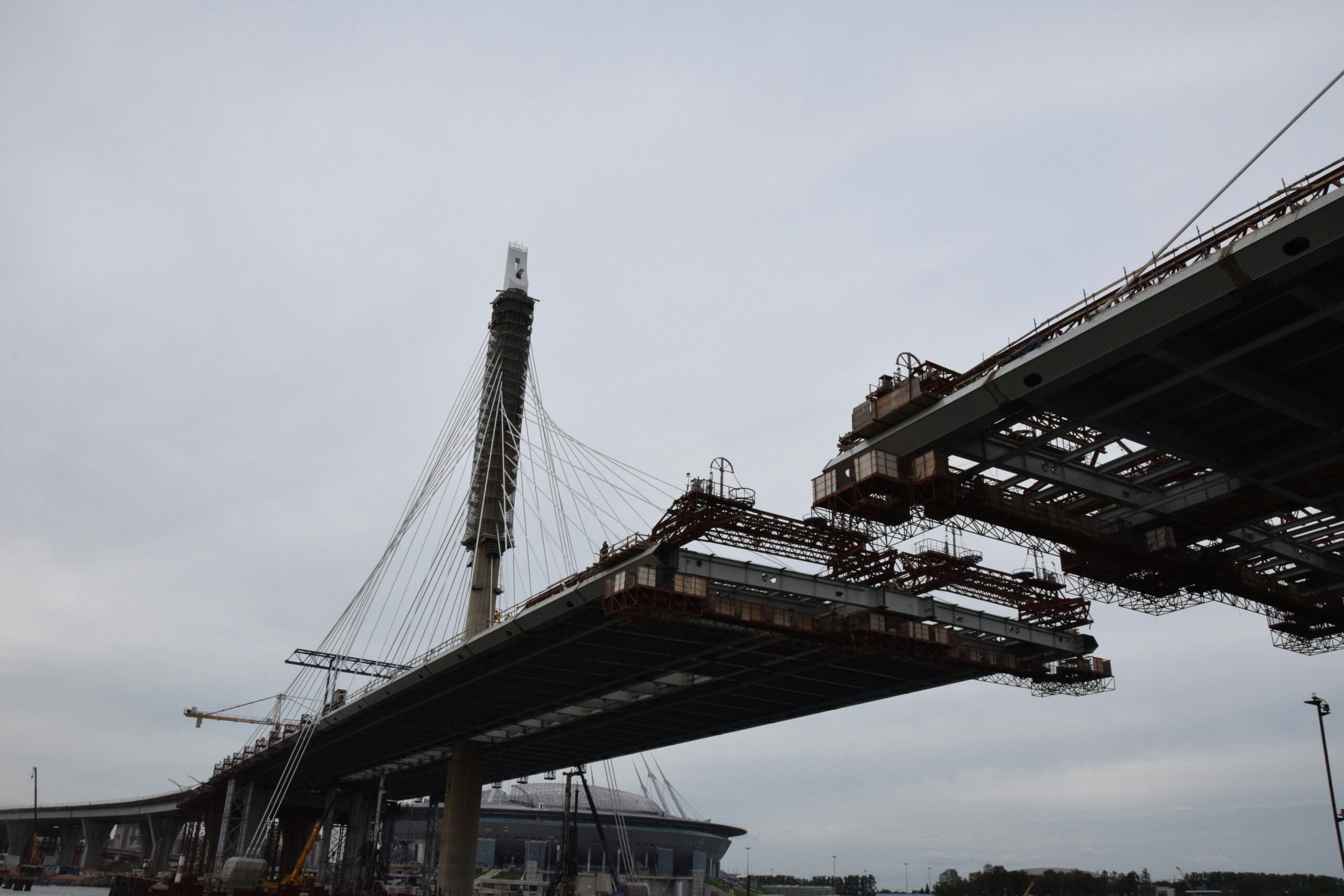 Under construction cable-stayed bridge with a tall support tower and visible steel framework against a cloudy sky.
