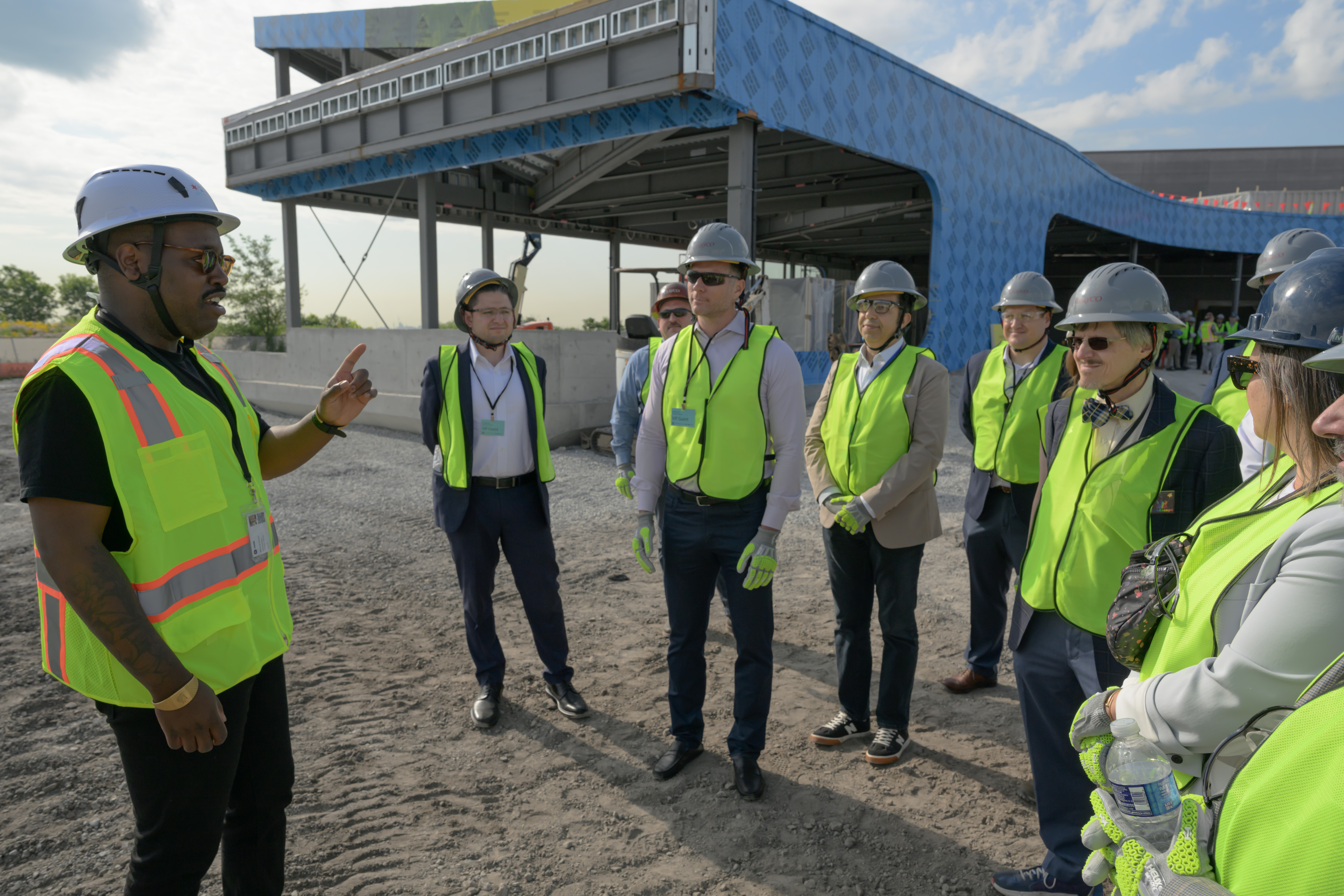 A man gestures to a group of people standing in front of a structure under construction. All wear yellow safety vests and white hard hats.